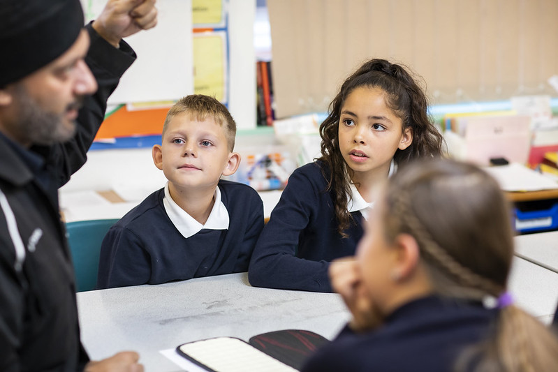 Three children look up at a volunteer sitting with them at their classroom table.