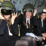 Smiling students sit in aircraft and try on flight helmets.