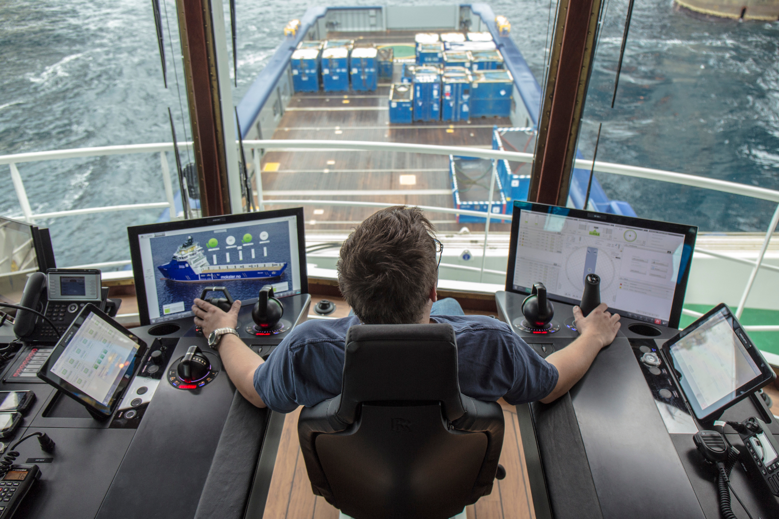 Person sits in a chair and holds controls. They are looking out onto the front of a ship. Either side of them is a screen displaying graphics and a diagram of a ship.
