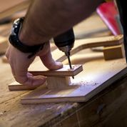 A hand holds down a piece of wood as a hole is drilled into it