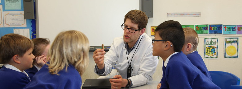 A volunteer wearing a white coat sits in the middle of a group of primary children who are looking at an object he holds