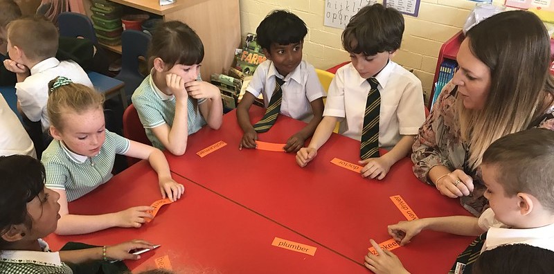 A group of primary children sit around a red classroom table with a volunteer. They each hold a piece of paper which says a different job role.