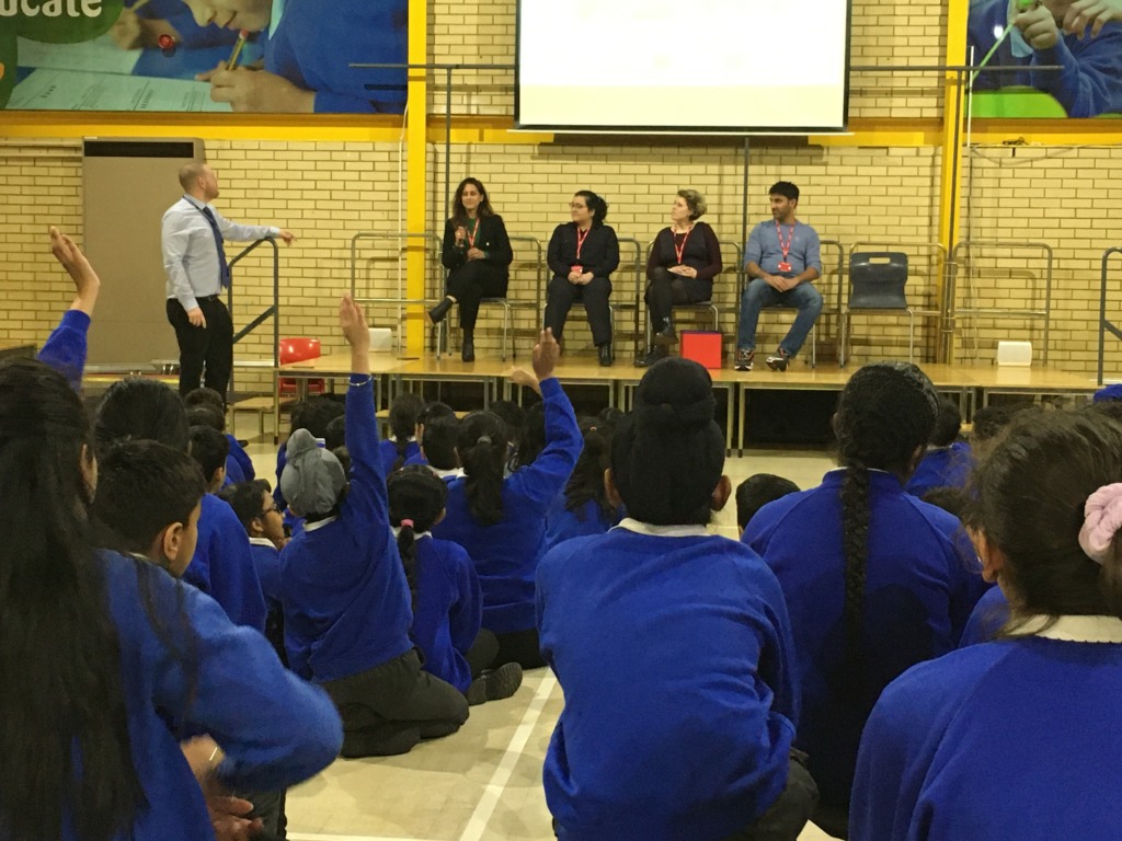 Children sit on the school hall floor wearing blue jumpers, several with their hands up. On the stage are four volunteers, and a teacher stands beside them.