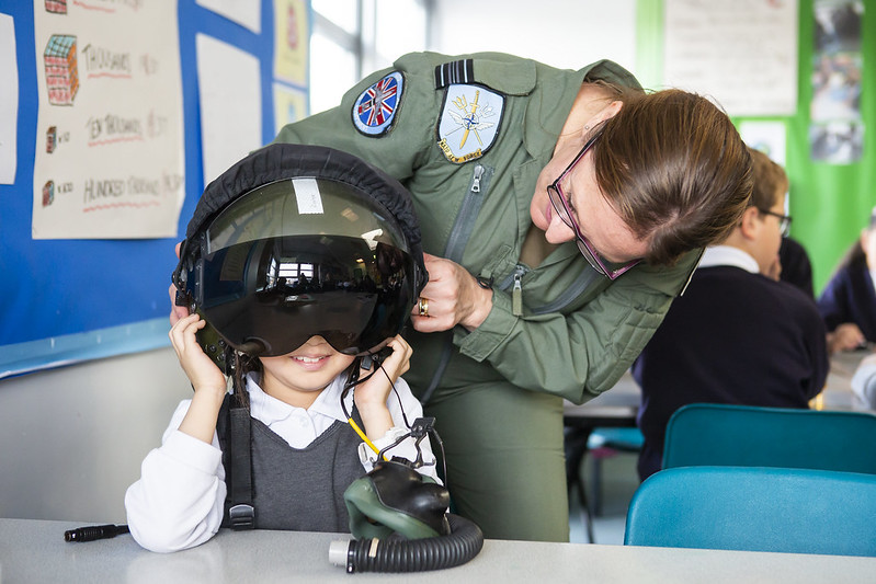 Female pilot in school with a young girl trying on her inflight helmet.