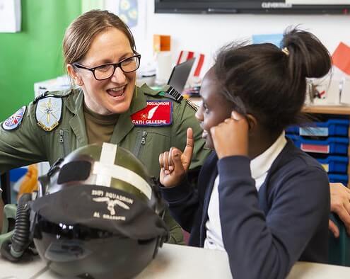 Primary aged girl chattging and laughing with a female RAF pilot in her uniform in her school classroom. 