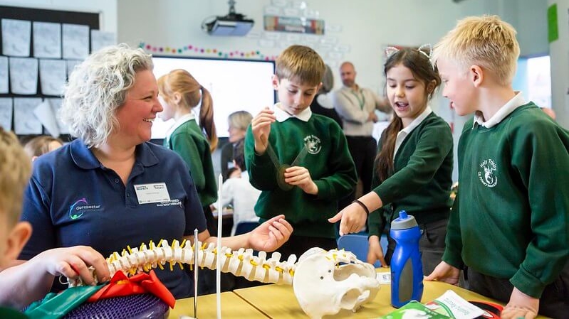 A female volunteer in a medical uniform sits with a small group of mixed primary children, a girl points at the model of a skeleton on the desk; the volunteer is laughing.