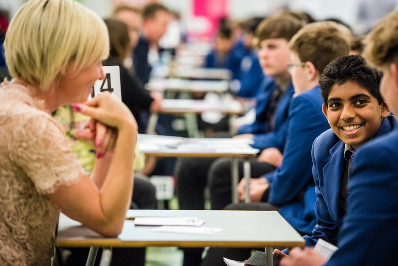 female volunteer doing speed networking with two secondary students at a small desk - other volunteers can be seen behind doing the same thing.